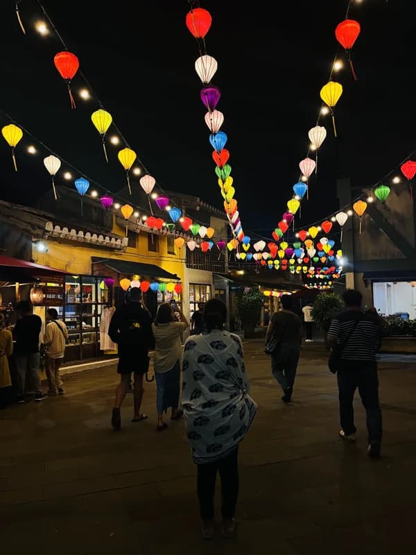 Travellers walk beneath colorful lanterns in an evening street scene.